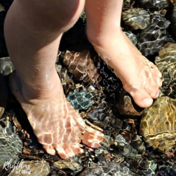 Child barefoot outside with feet in the water standing on river stones.