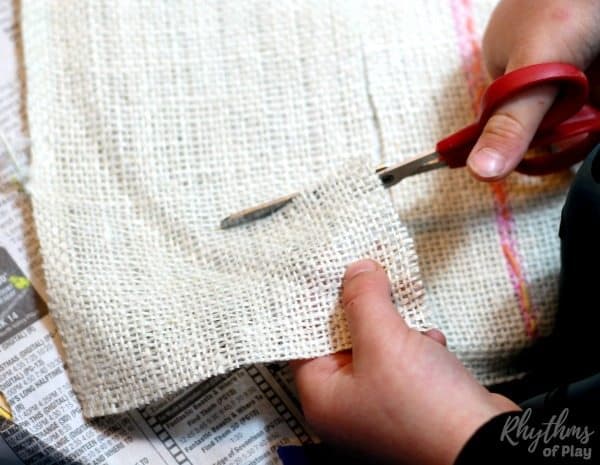 Kid cutting a square of burlap (or another fabric) to swaddle a small peg doll to make Baby Jesus craft.