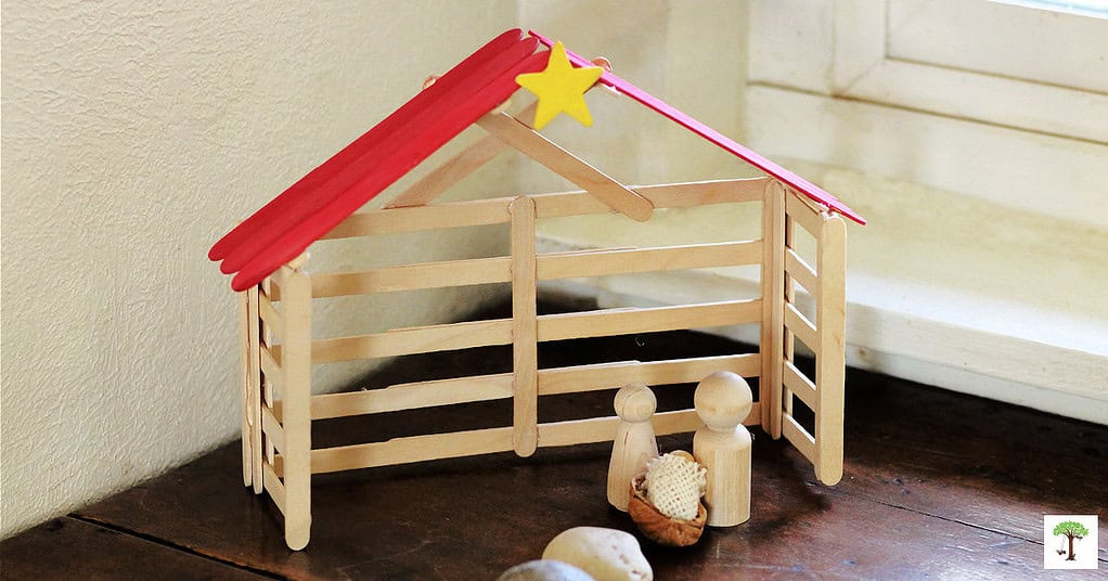 Popsicle stick Nativity stable Christmas craft for kids and adults with Mary, Joseph, and Baby Jesus sitting underneath the red jumbo stick roof.