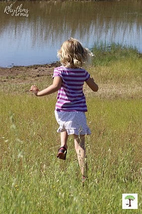 Young girl running through tall grass beside a pond enjoying playtime in nature.